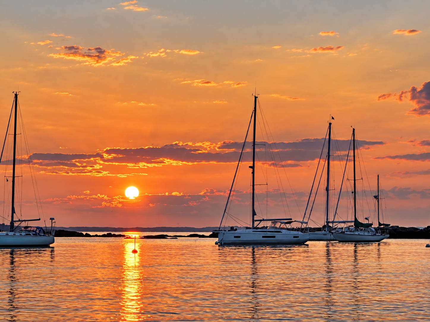 Sailboats on their moorings at sunset, the sun low over the breakwater